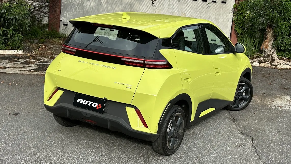 BYD Dolphin Mini amarelo parado de traseira em frente a um portão branco com árvores ao fundo