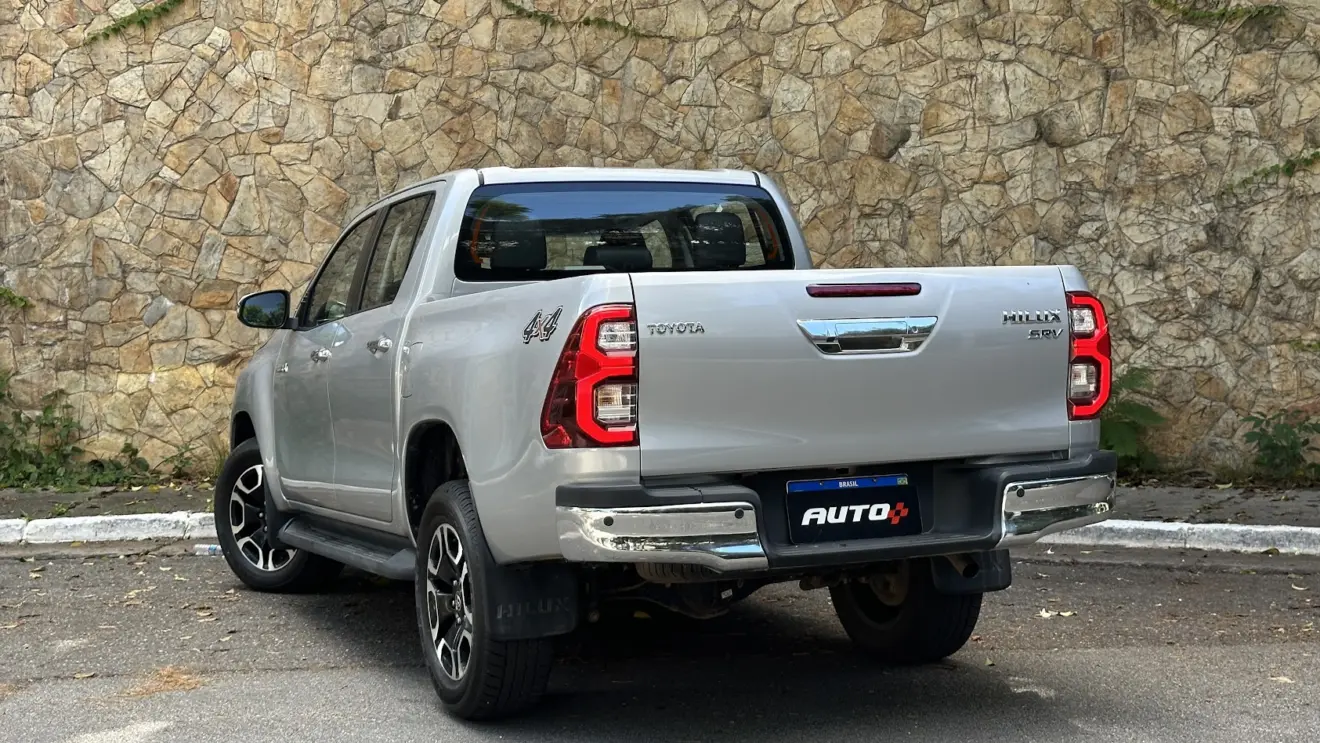 Caminhonete  Toyota Hilux SRV prata estacionada de traseira com muro de pedras ao fundo