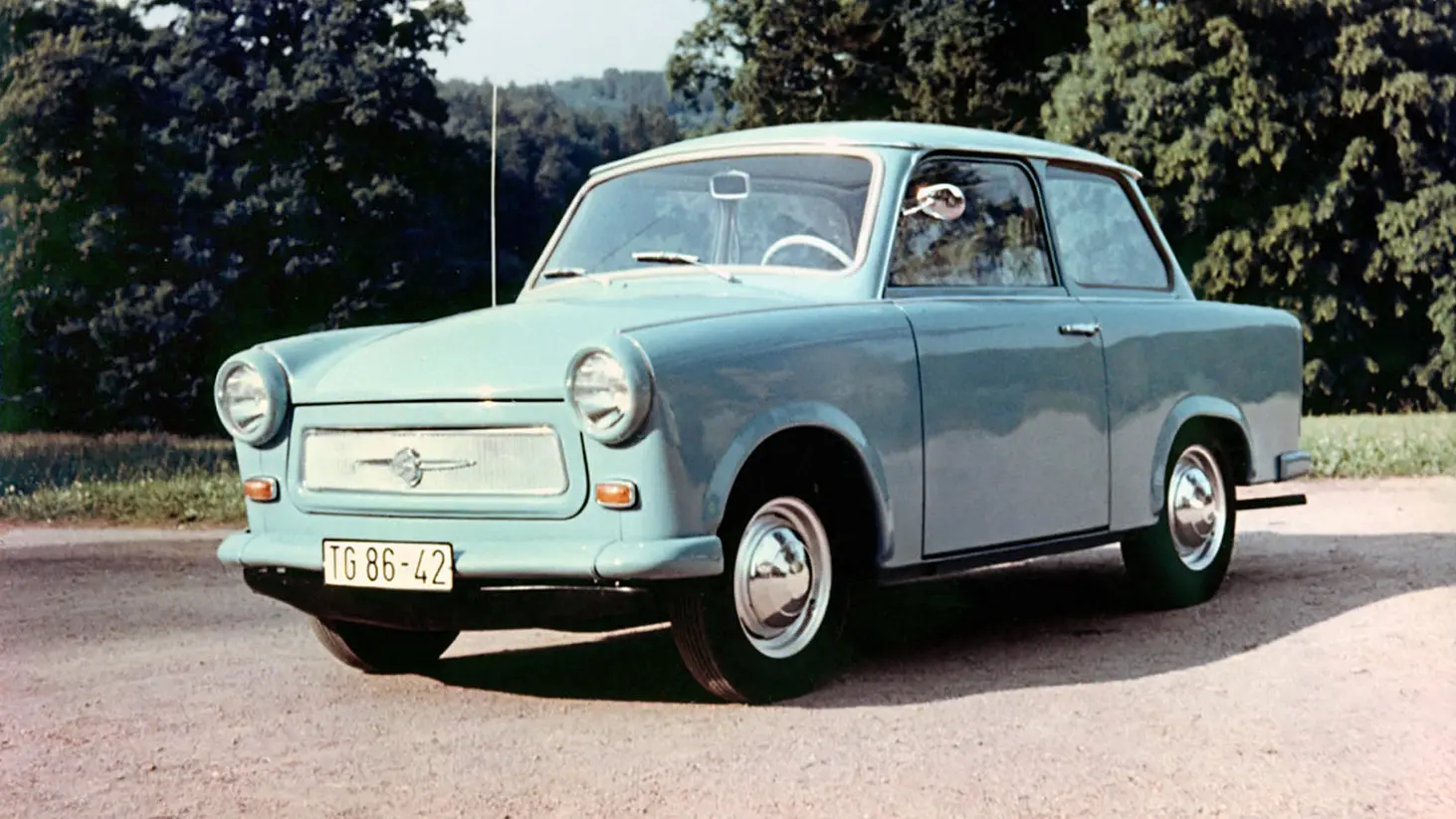 Trabant 601 azul parado de frente no asfalto com árvores ao fundo