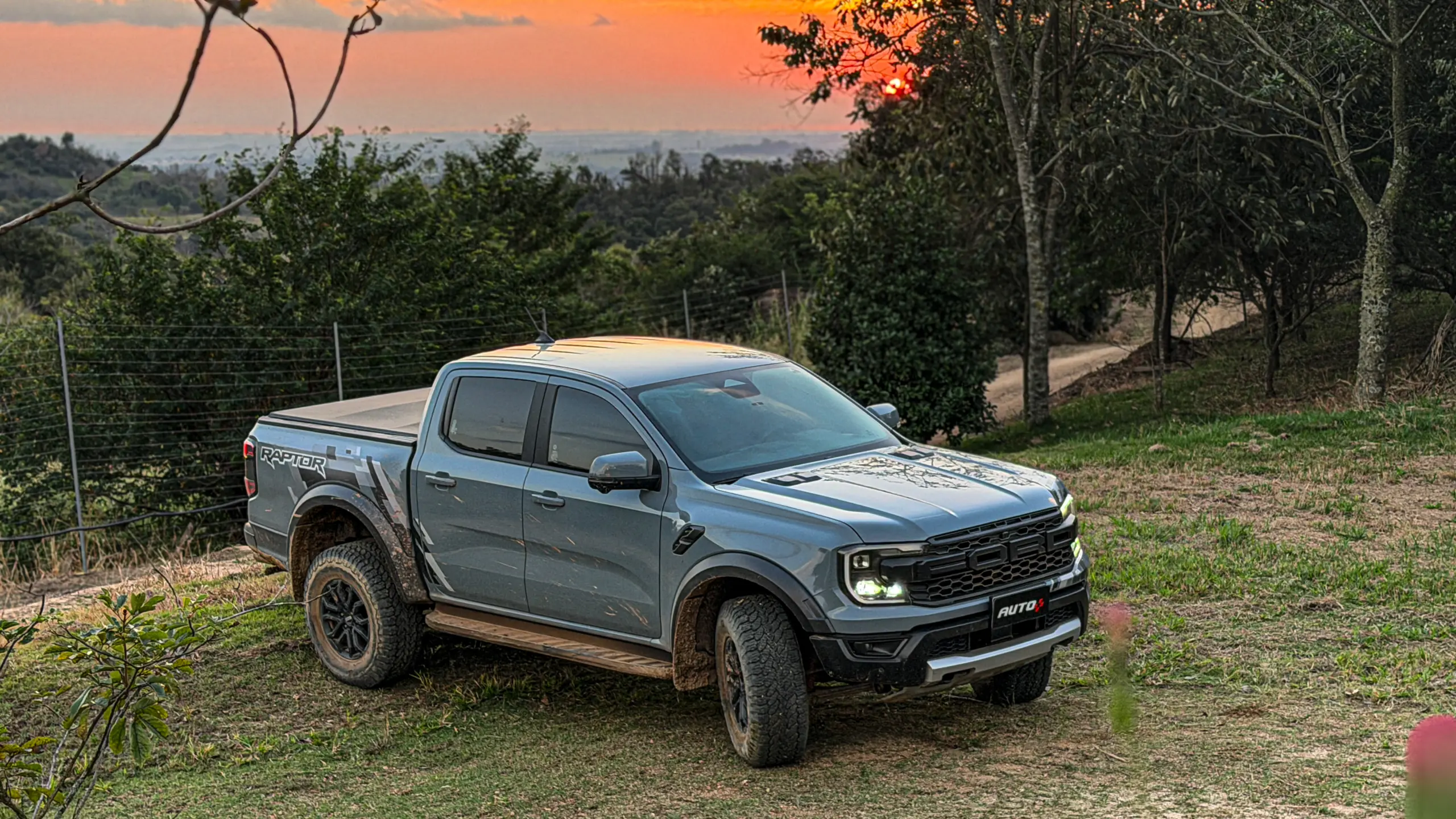 Ford Ranger Raptor suja de barro e terra estática na grama na cor Cinza Interlagos com pôr do sol